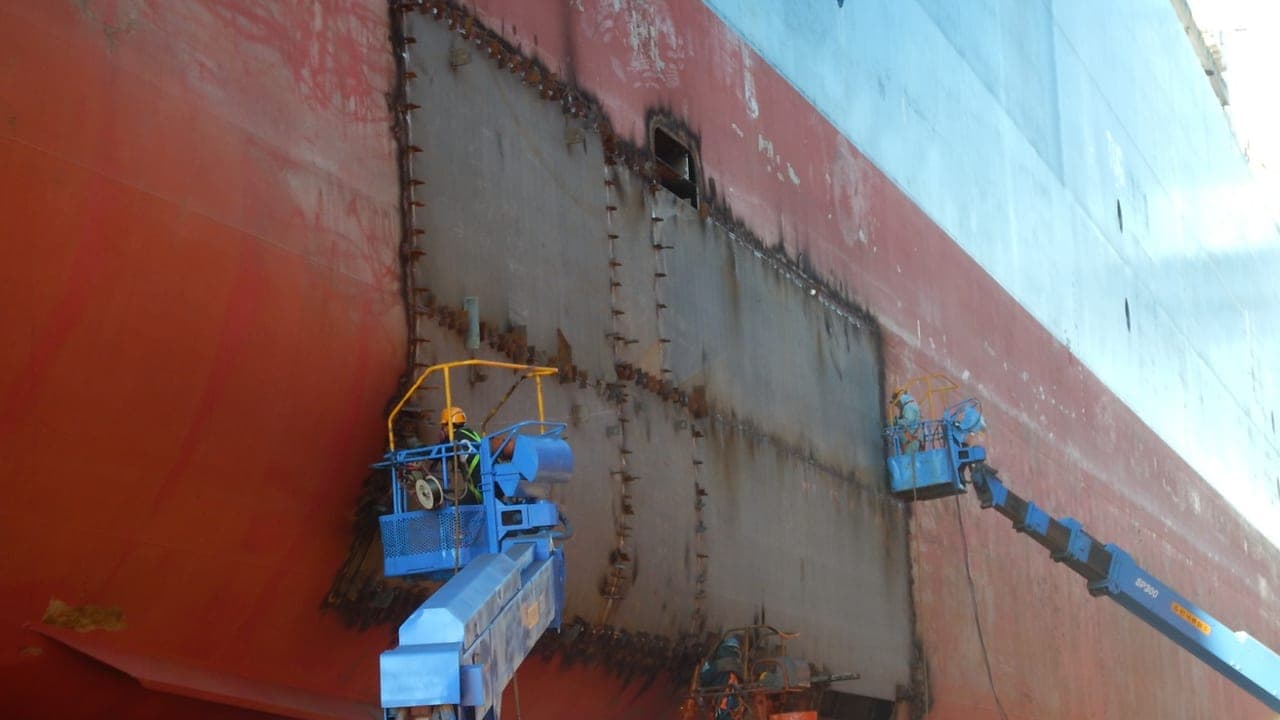 Shell plating cut out and renewed in drydock, with welders working from cherry pickers alongside the hull.