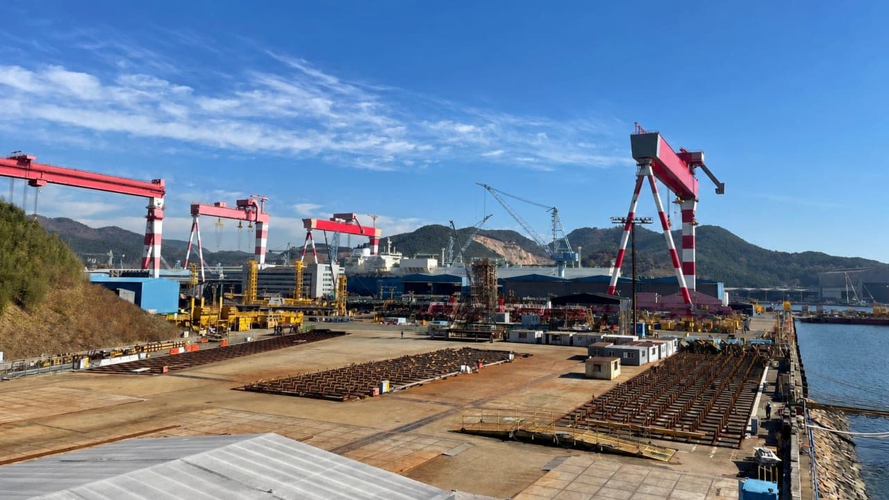 A shipyard building berth under red and white gantry cranes, with steel sections laid out across the apron.